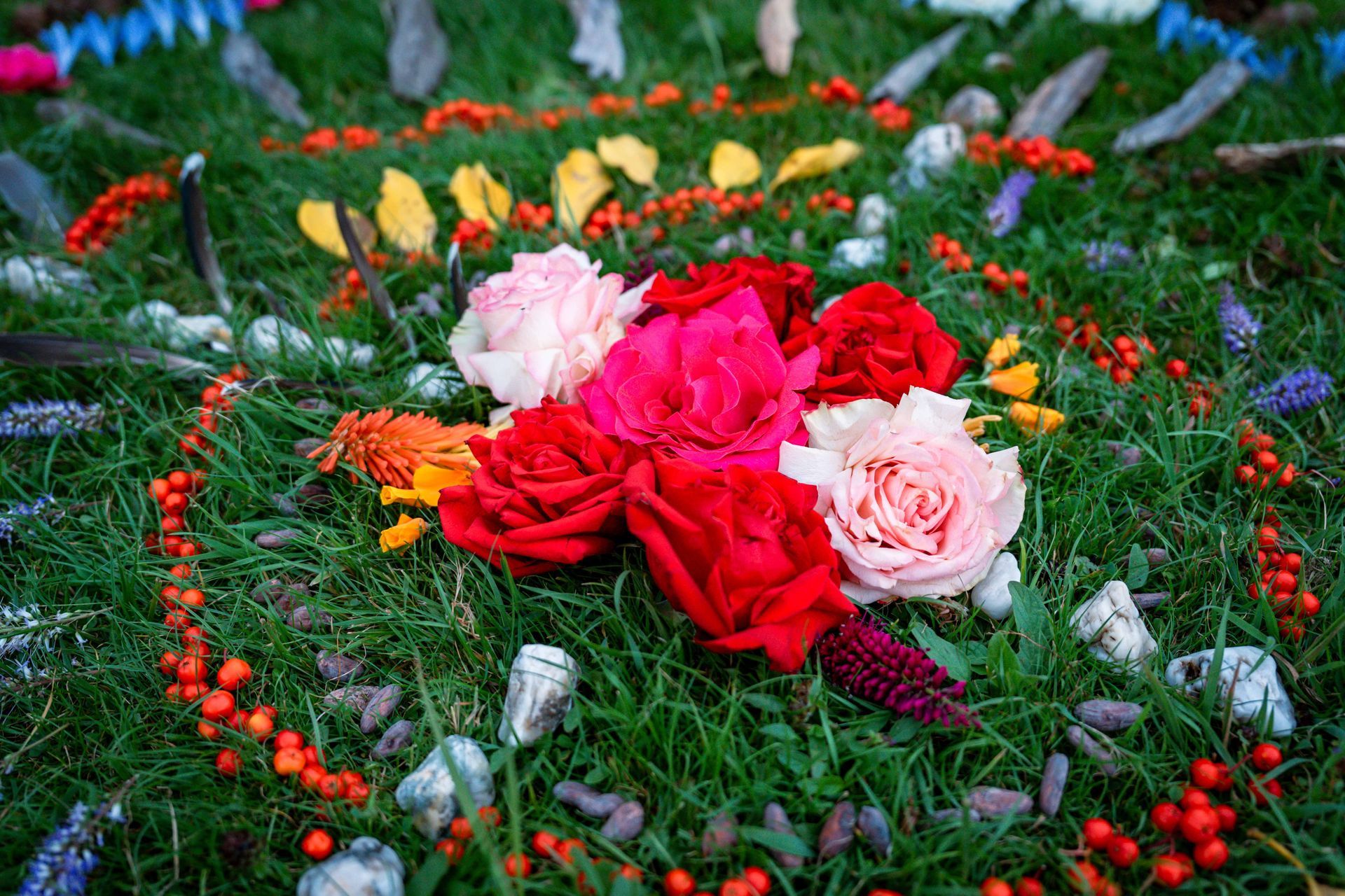 A circular flower arrangement on grass, with red, pink, and orange blooms surrounded by berries and feathers.