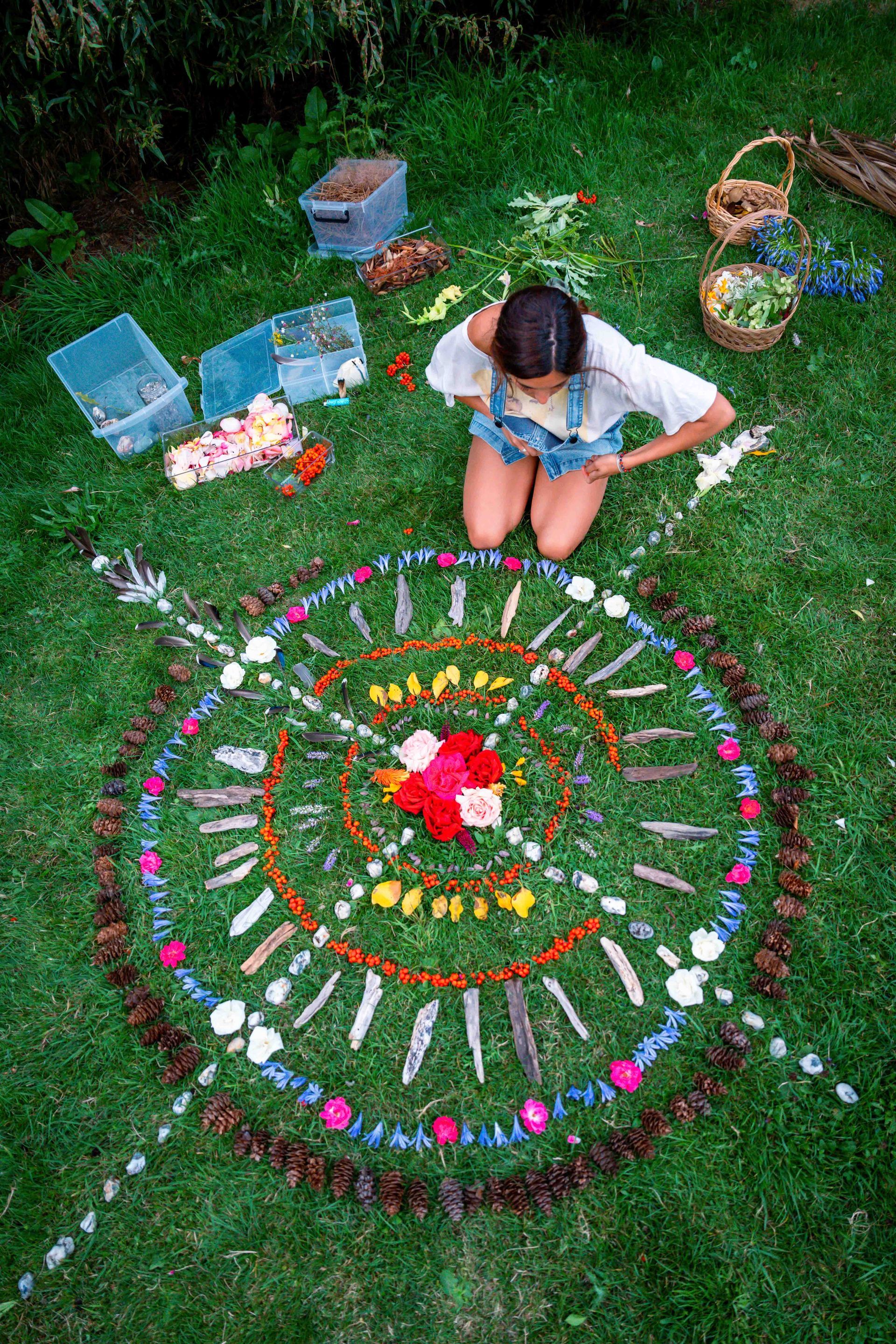 Person arranging flowers and natural items in a circular mandala on grass, outdoors.