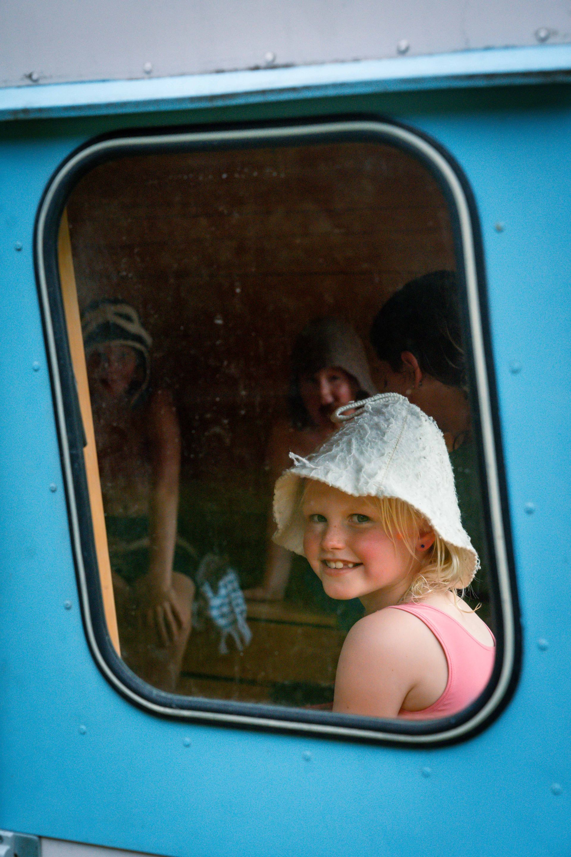 Girl in a bucket hat smiles from a blue-framed window. Other figures are inside a wooden room.