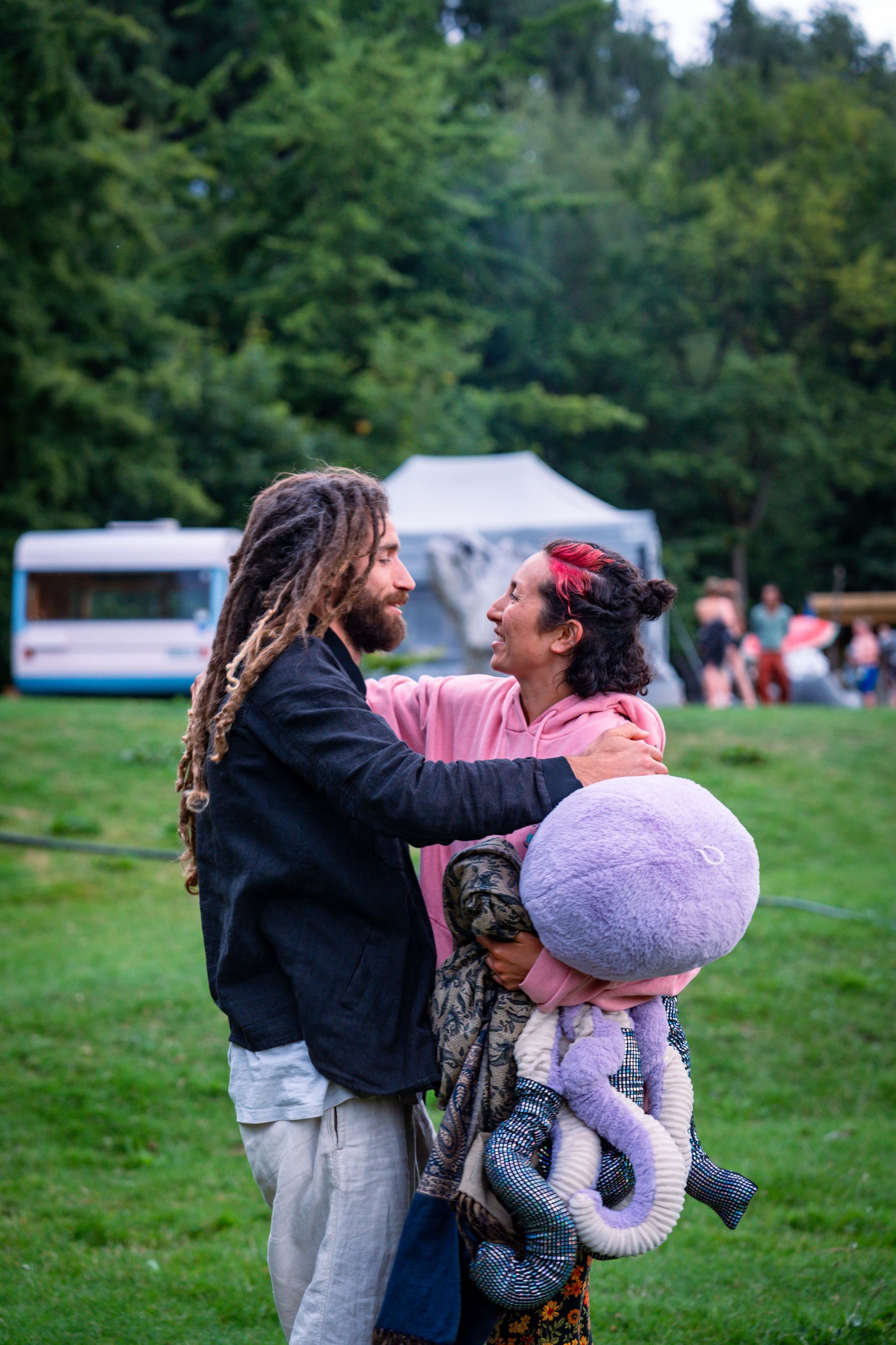 Man with dreadlocks hugs smiling person holding plush toys in a field.