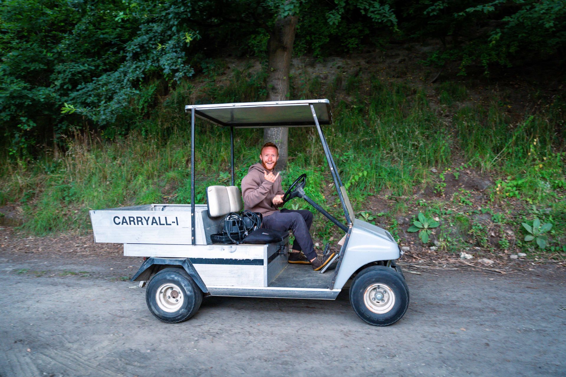 Person in golf cart, smiling, giving thumbs-up. Cart is on a dirt path, with trees in the background.
