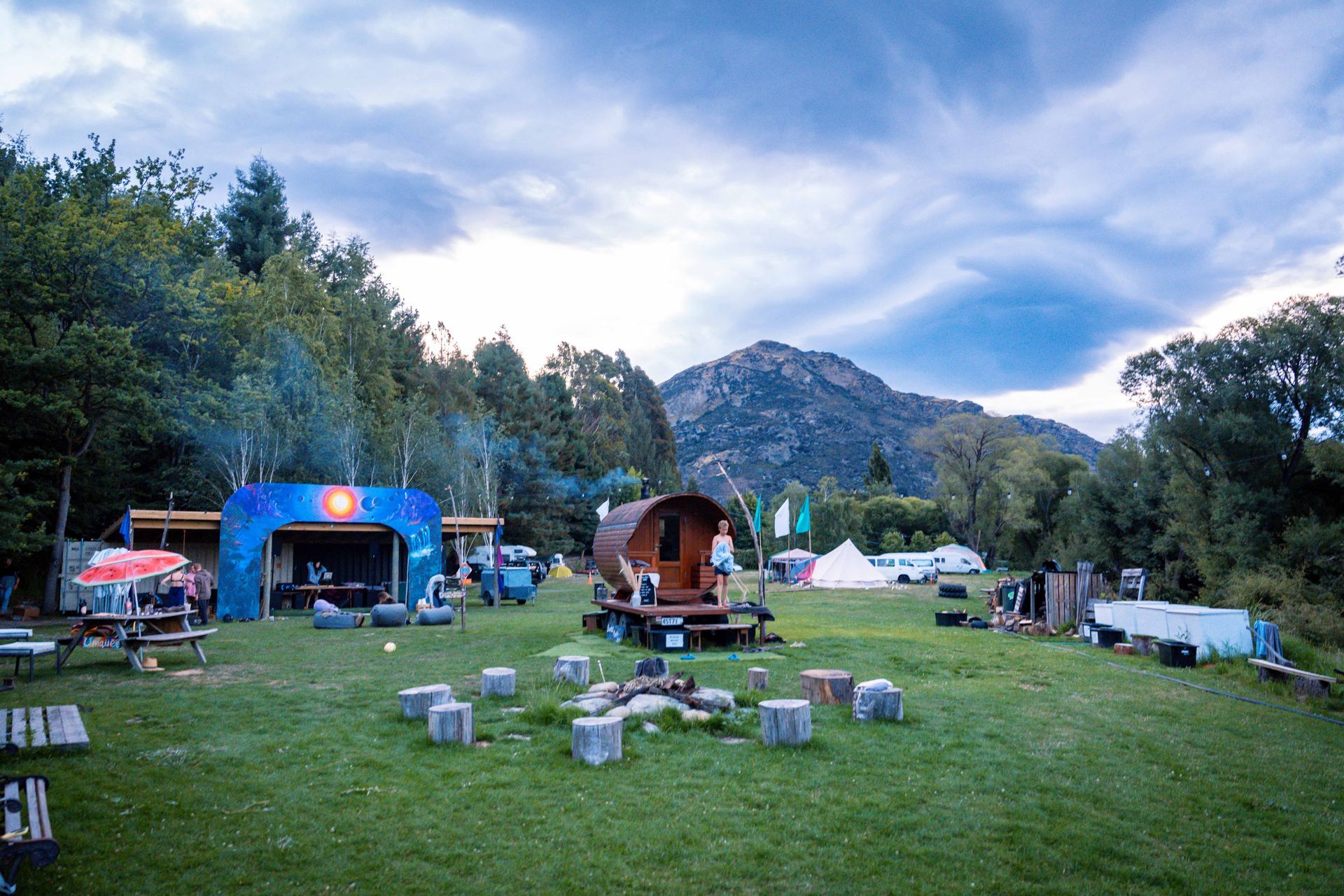 Camping area with a wooden wagon, stage, fire pit, and mountain backdrop under a cloudy sky.