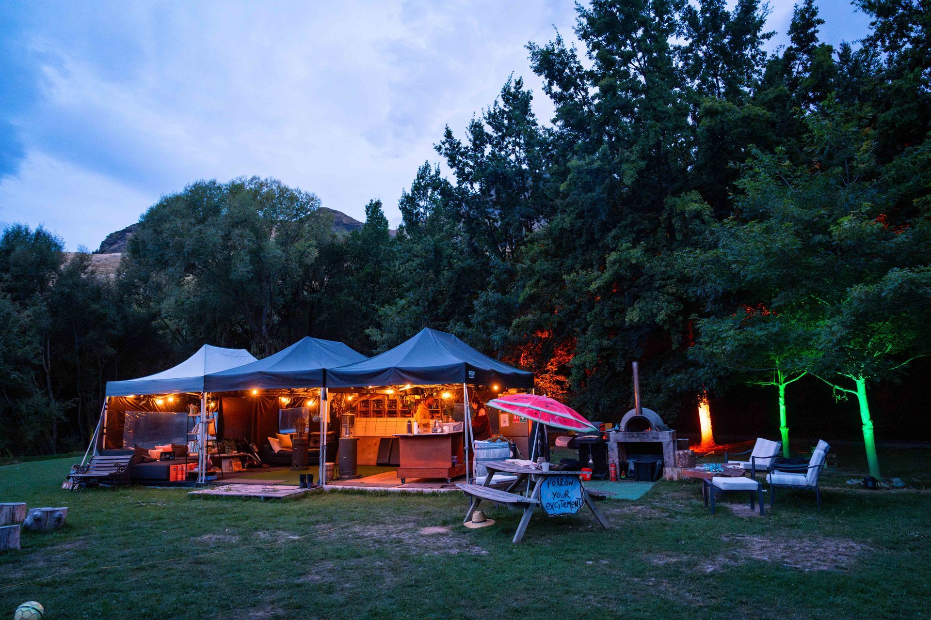 Outdoor gathering space at dusk, string lights, tent, pizza oven, trees, seating.