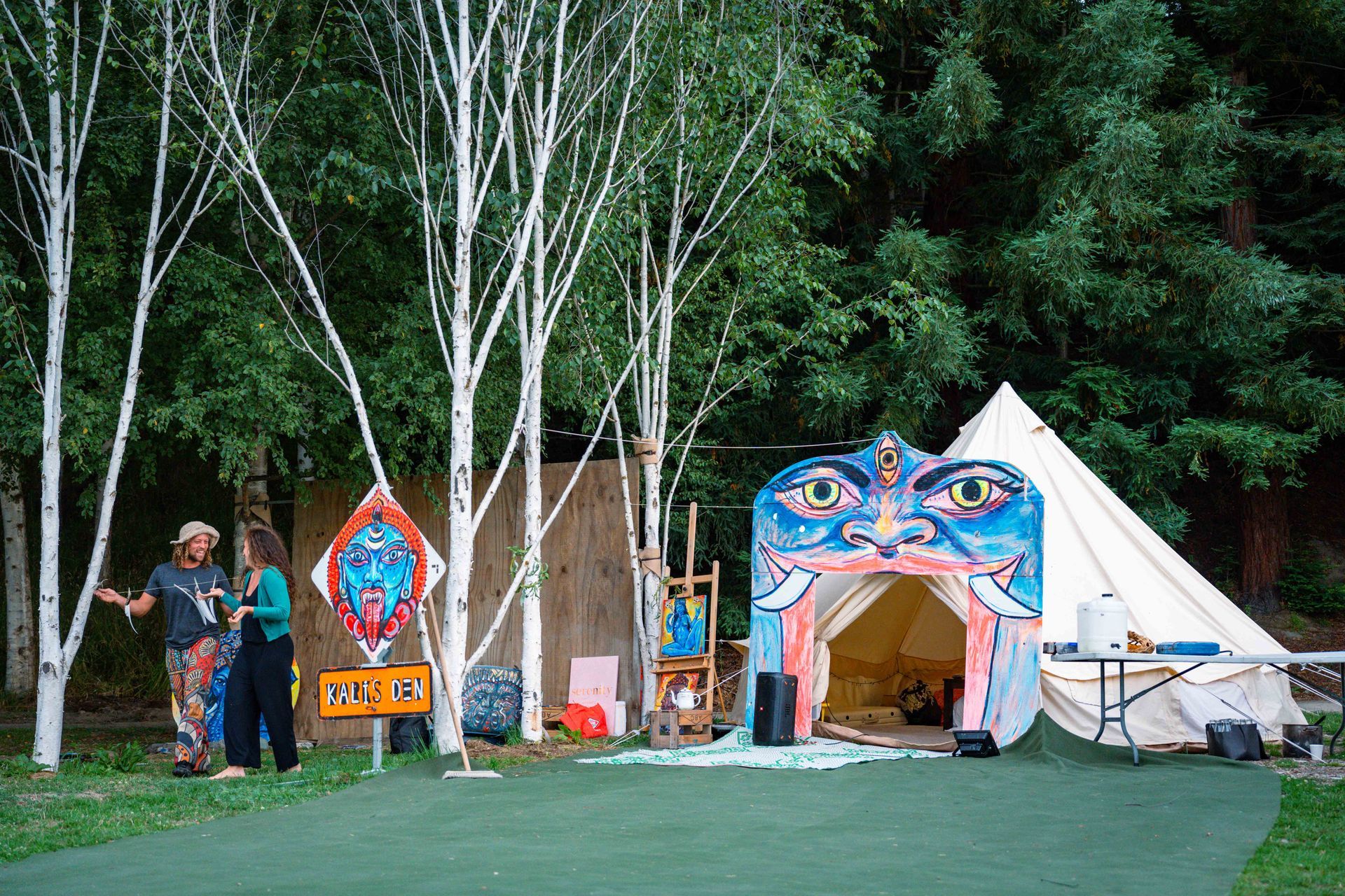 Outdoor event with art, tents, trees, and people; colorful art piece, white-trunked trees, green grass area.
