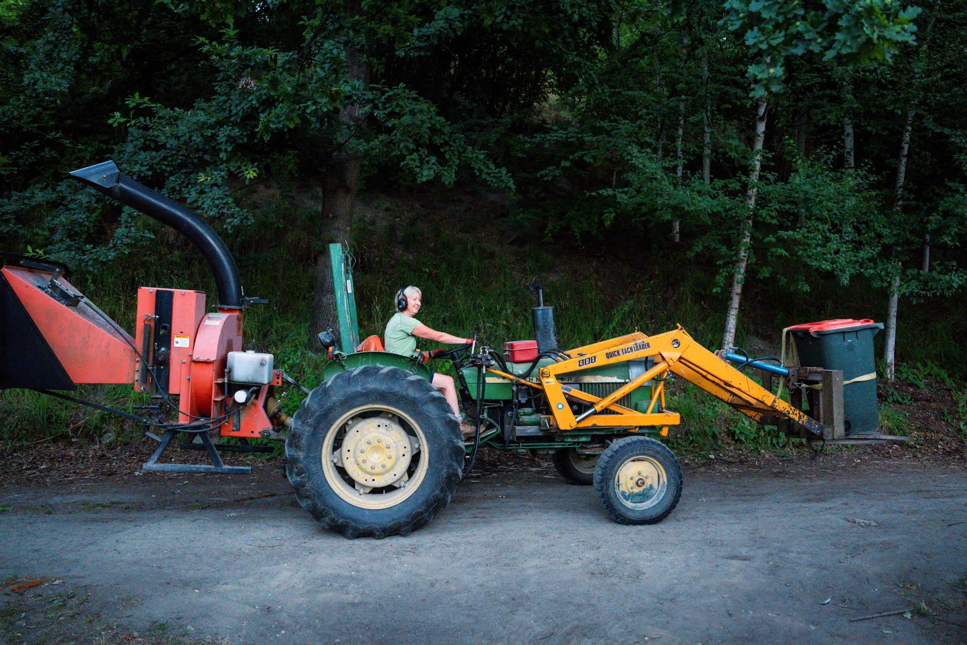 Man operating a tractor with a wood chipper and trash bin in a wooded area.