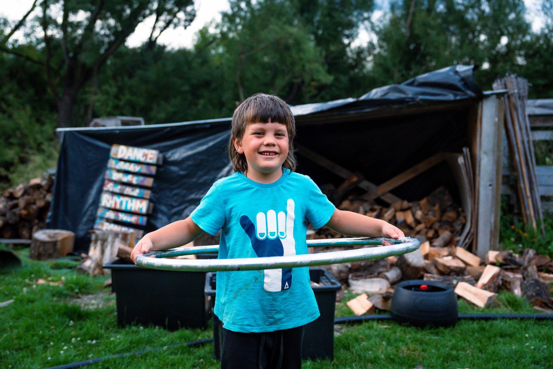 Boy with brown hair smiles, holding a hula hoop. He's wearing a blue shirt. Outdoors with firewood and shed in background.