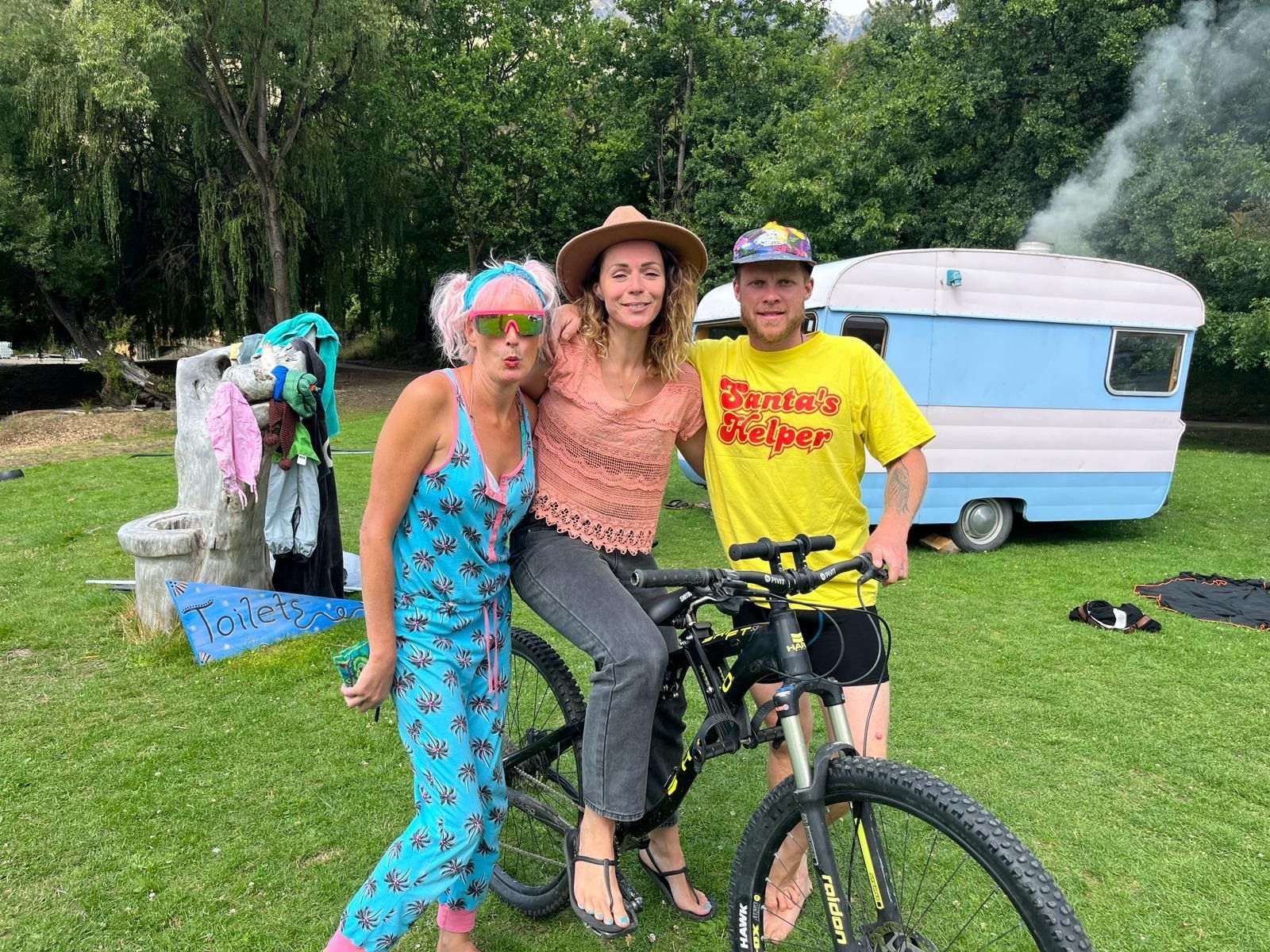 Three people pose with a bicycle in front of a blue and white camper, on a green lawn.