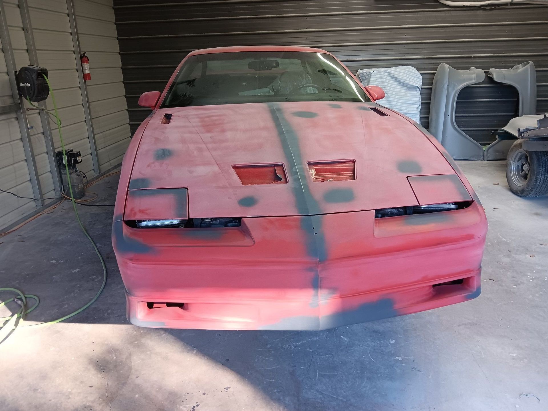A red Pontiac Firebird undergoing bodywork and primer in a garage, with a matching bumper panel resting in the background.