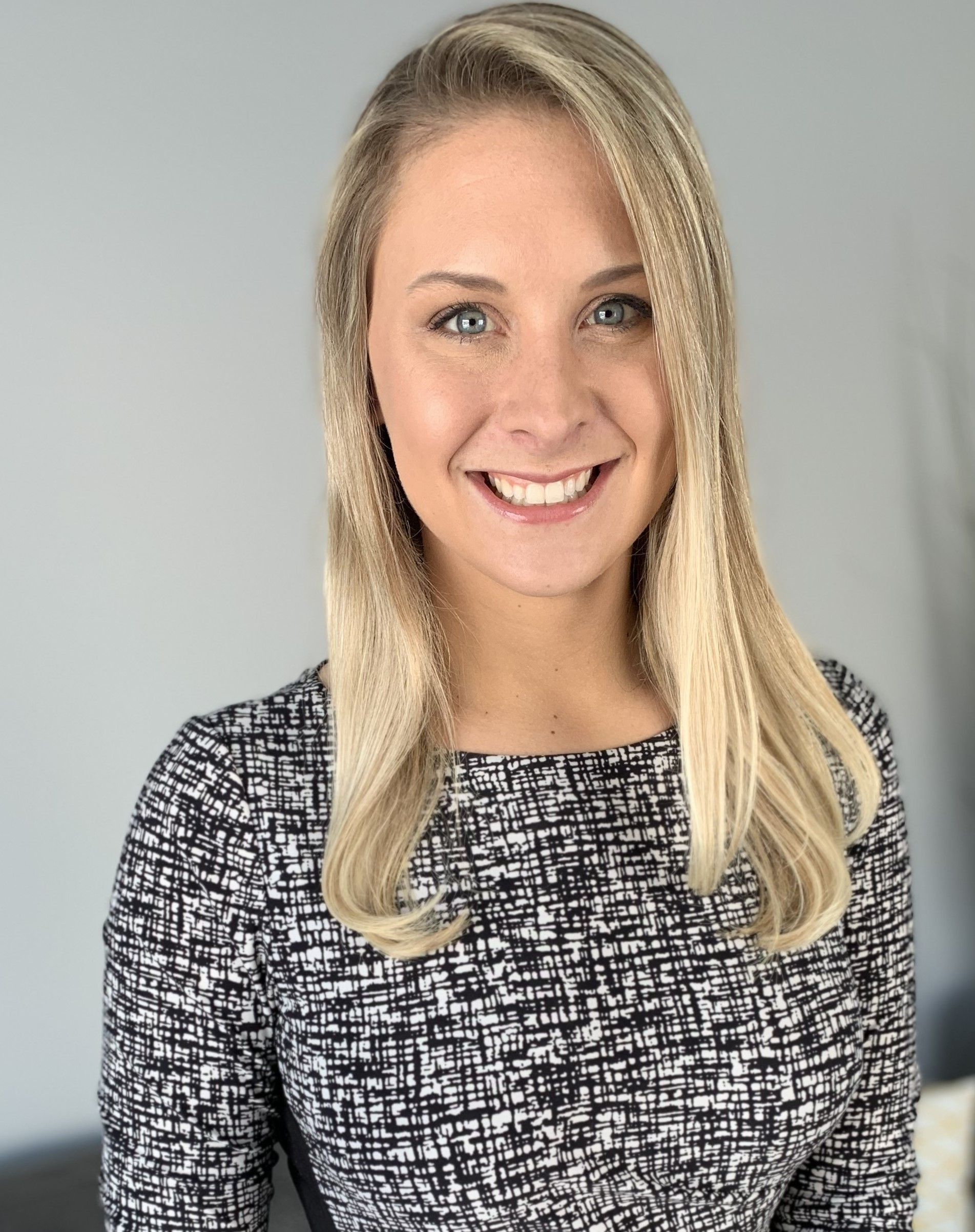 A smiling woman with blonde hair, wearing a black and white patterned top against a plain gray background.