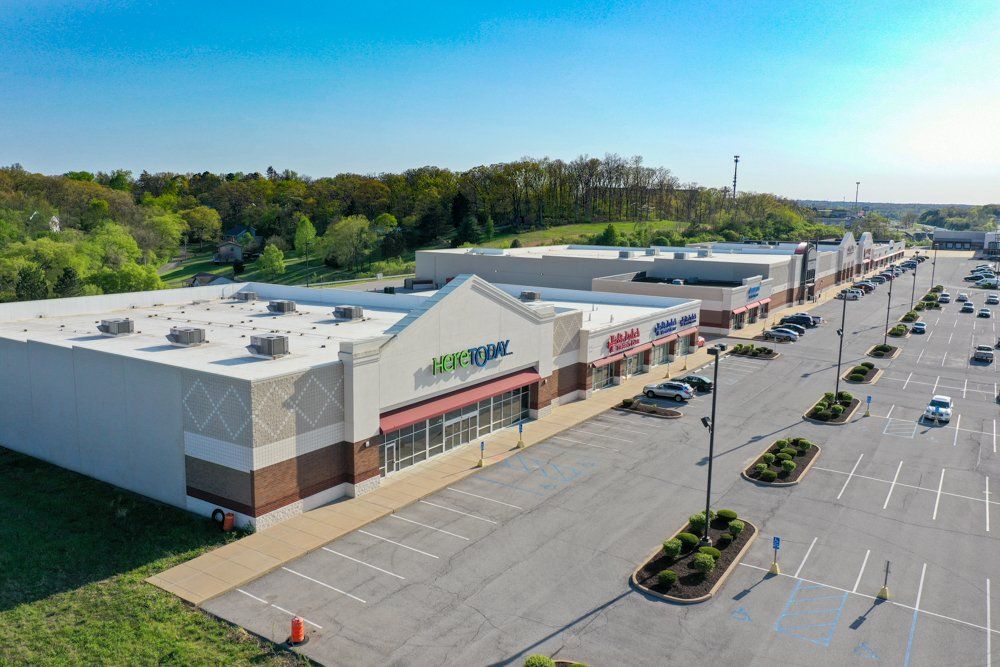 Shopping center with stores, parking lot, and trees under a blue sky.