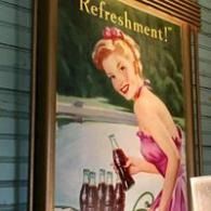 Retro soda ad poster with a smiling woman in a pink dress holding a bottle under the word “Refreshment!”