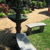 Garden fountain beside a stone bench on a gravel path, surrounded by greenery and flowers