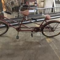 Rusty red tandem-style bicycle with brown seats parked on a sidewalk indoors.