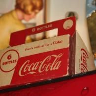 Red Coca-Cola six-pack carton on a counter, with blurred people in the background