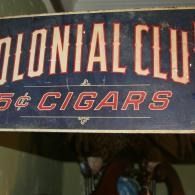 Blue sign reading “Colonial Club 5¢ Cigars” in cream and red lettering