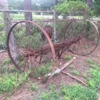 Rusty old farm machinery with large wheels in a grassy outdoor area near a fence.