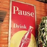 Red sign reading “Pause Drink” with a soda bottle illustration on a wooden wall
