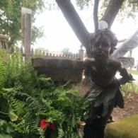 Small garden statue beside lush ferns and a red flower under a tree