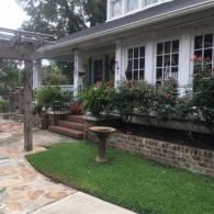 Front porch of a white house with brick steps, potted plants, and a green lawn.