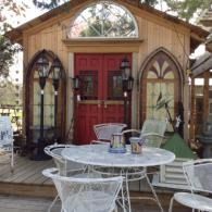 Rustic garden shed with red double doors and arched windows, fronted by a white metal patio table and chairs.