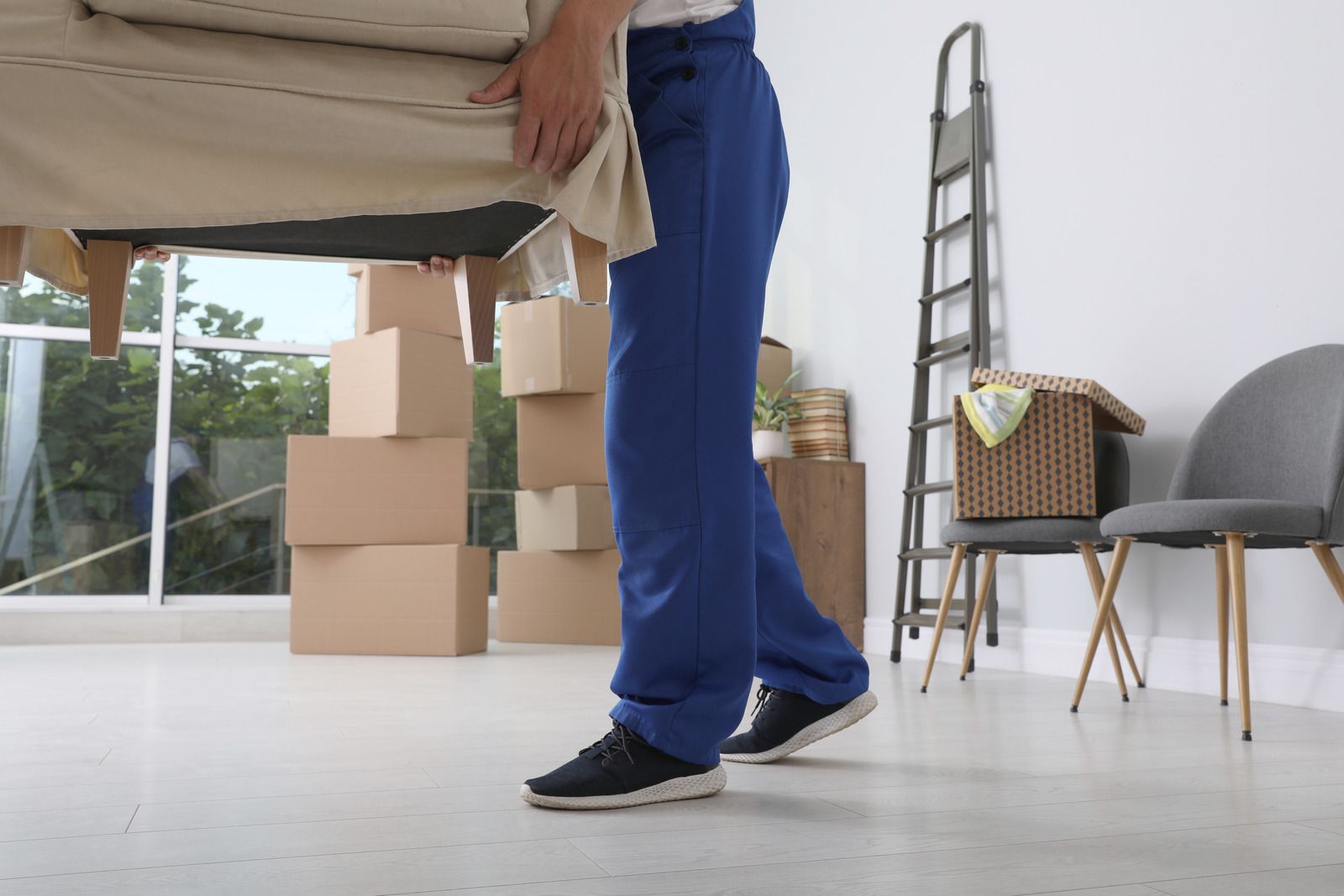 Person in blue pants lifting a sofa, moving boxes in a bright room.
