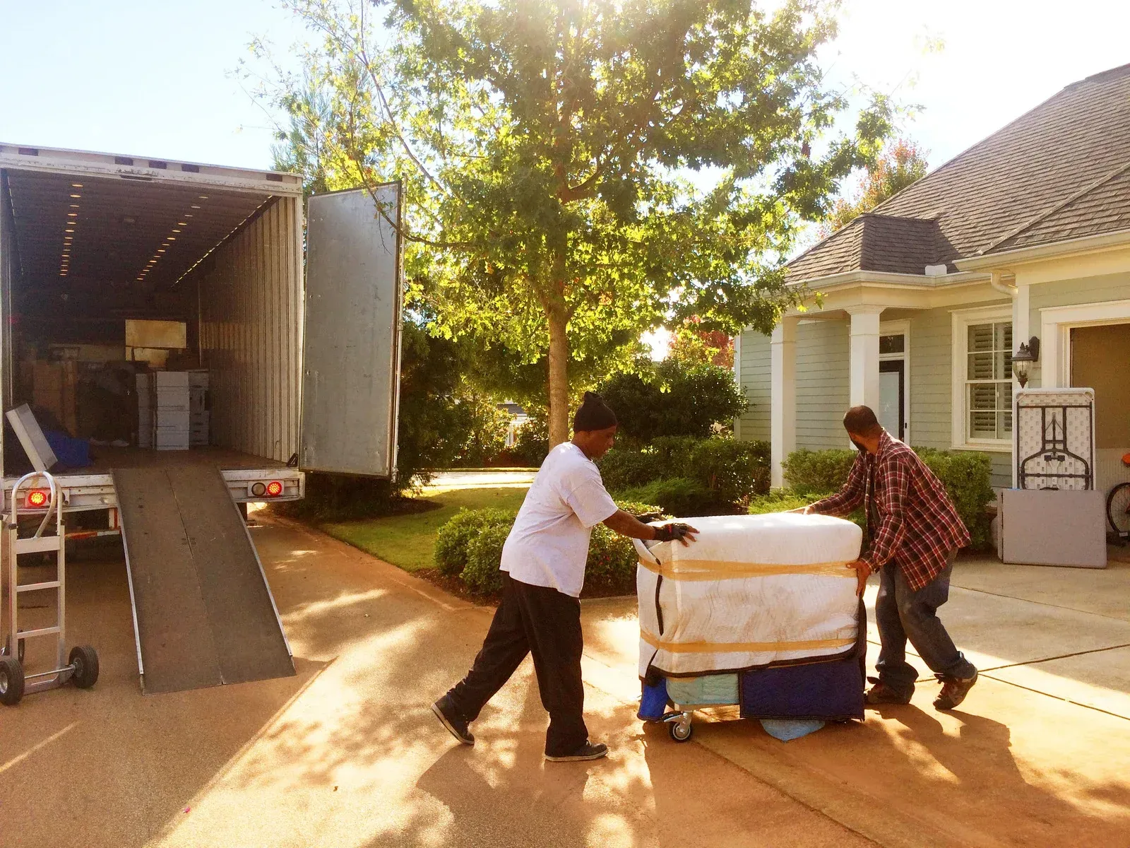 Two people pushing a moving cart with furniture toward a truck ramp in front of a house. Sunny day.