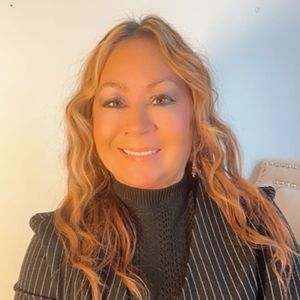 A person with long blonde wavy hair, wearing a black pinstriped blazer over a dark turtleneck, smiles at the camera.