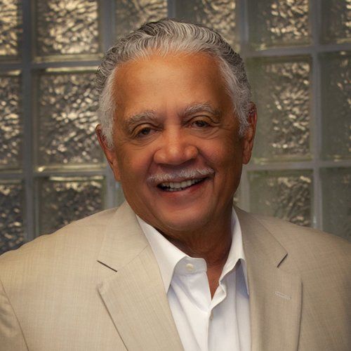 A smiling person with grey hair and a mustache wearing a beige suit and white shirt, posed against a glass block wall.