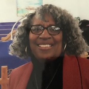 A smiling person wearing glasses and a red jacket, standing in what appears to be a church or event space.