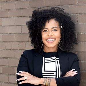 A smiling person with curly hair, wearing a black blazer over a geometric shirt, standing against a brick wall.