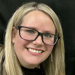 A smiling person with blonde hair and glasses wearing thin, vertical gold earrings against a black background.