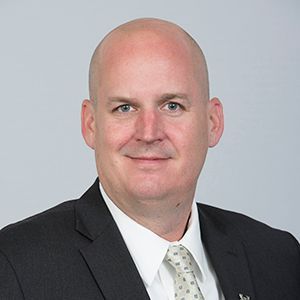 A person with a shaved head wearing a dark suit, white dress shirt, and a light-colored patterned tie against a grey wall.