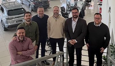 Sete homens posando em uma escadaria de uma concessionária de automóveis, provavelmente para uma foto de grupo.