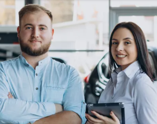 Homem e mulher sorrindo em uma concessionária de carros; o homem está com os braços cruzados. A mulher segura um tablet.