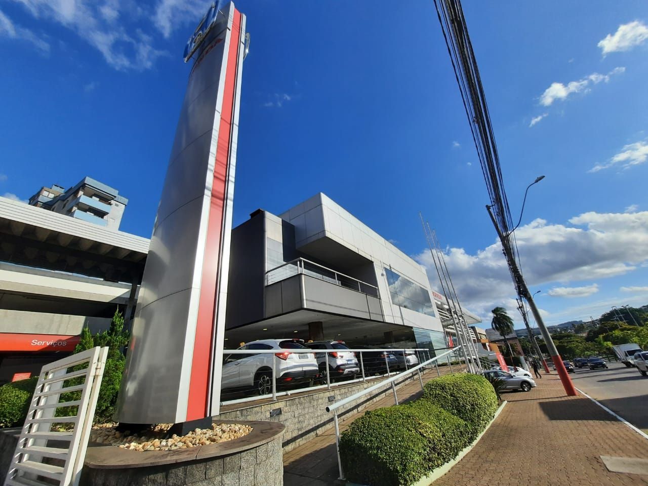 Fachada de concessionária de veículos com placa alta, carros em exposição e céu azul.
