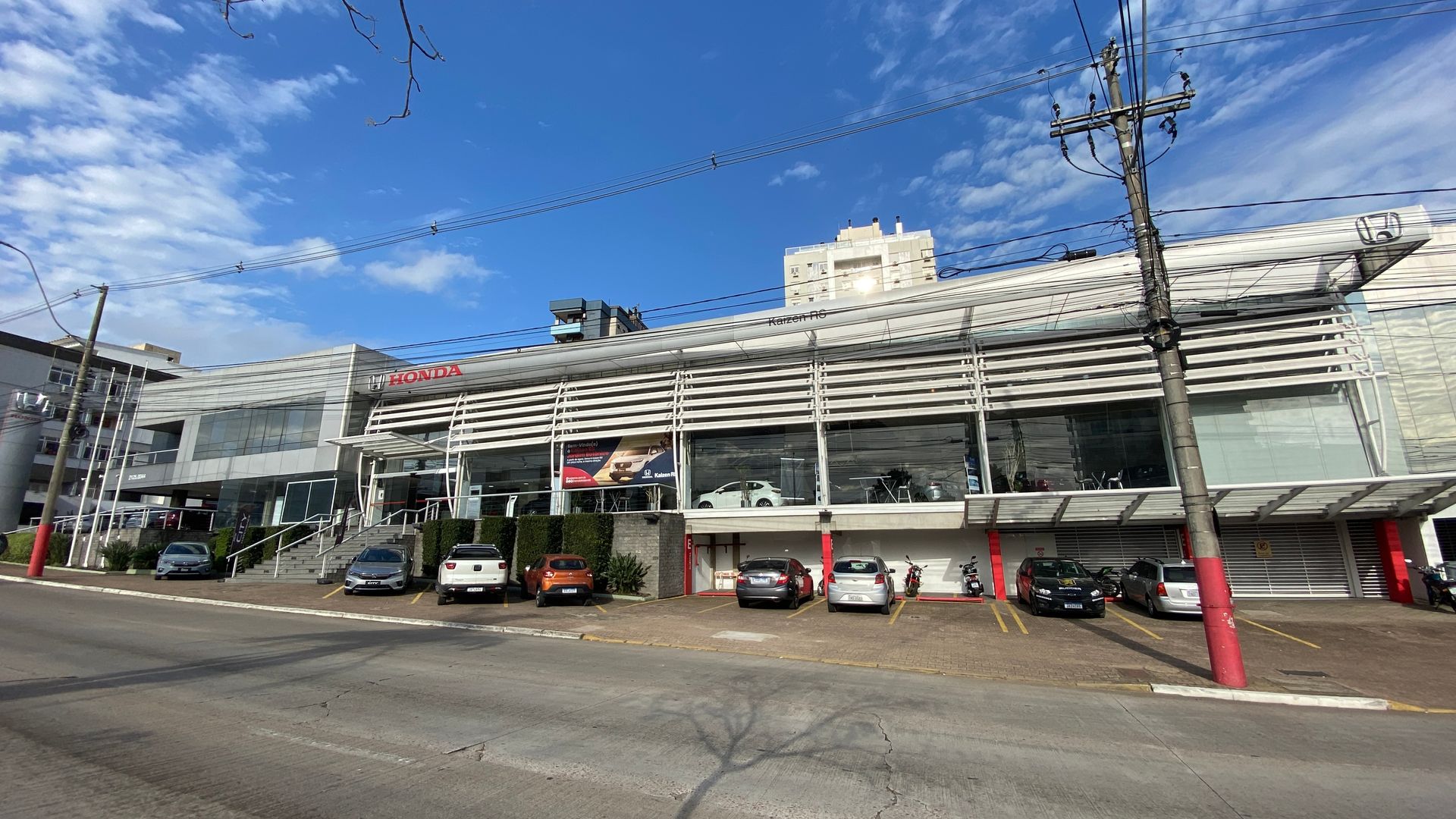 Fachada de um prédio, concessionária de automóveis. Carros estacionados do lado de fora. Céu azul.