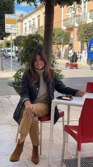 Donna seduta su gradini di pietra illuminati dal sole in riva al mare, indossa una camicetta chiara e sandali neri con una borsa di paglia.