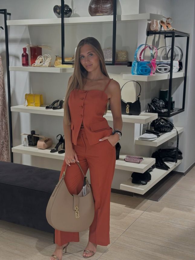 Una donna in piedi accanto a un muretto di pietra sul lungomare, con indosso una blusa colorata e jeans, sullo sfondo un lungomare storico.