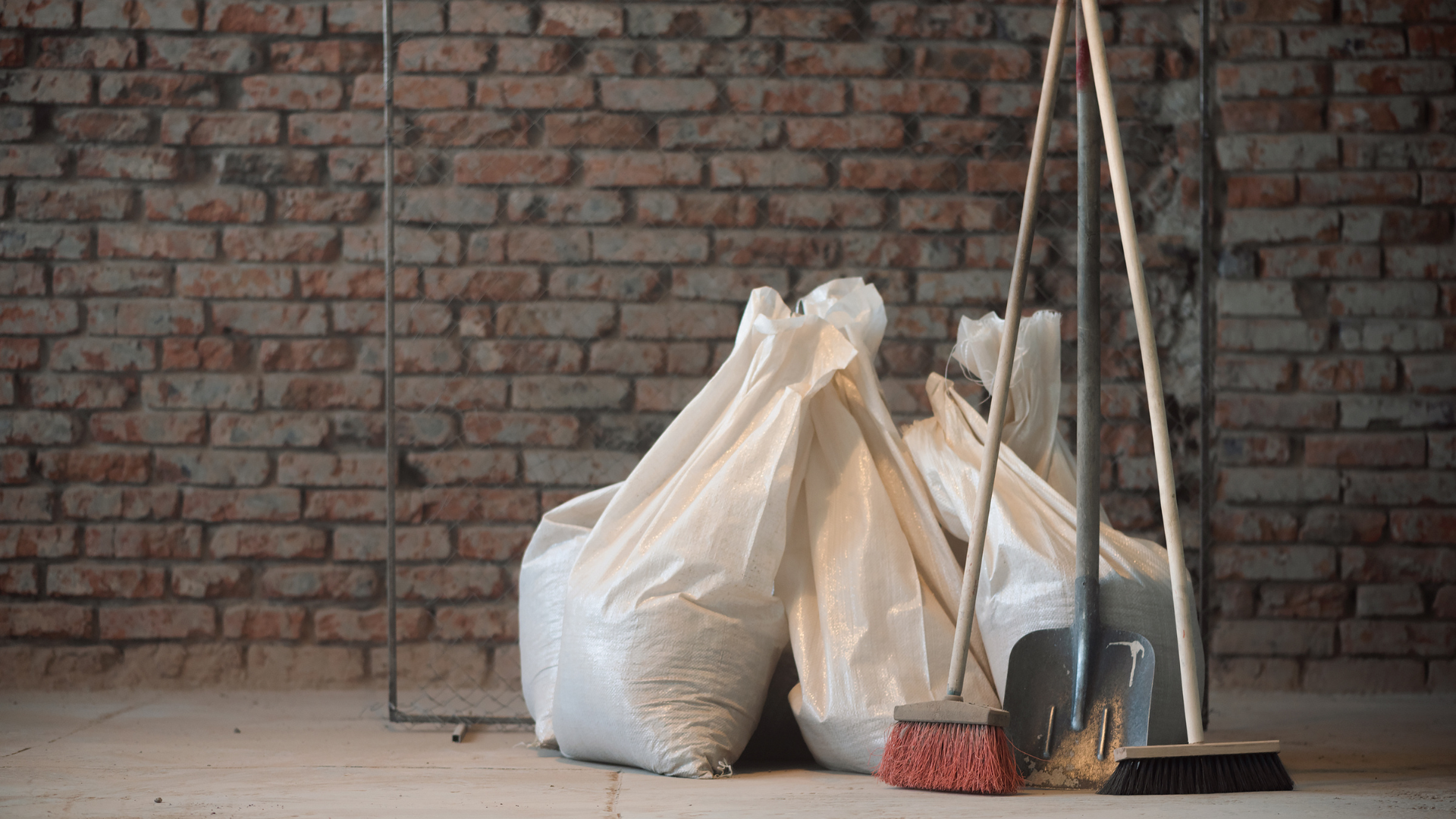 A pile of bags , a broom , and a shovel in front of a brick wall.