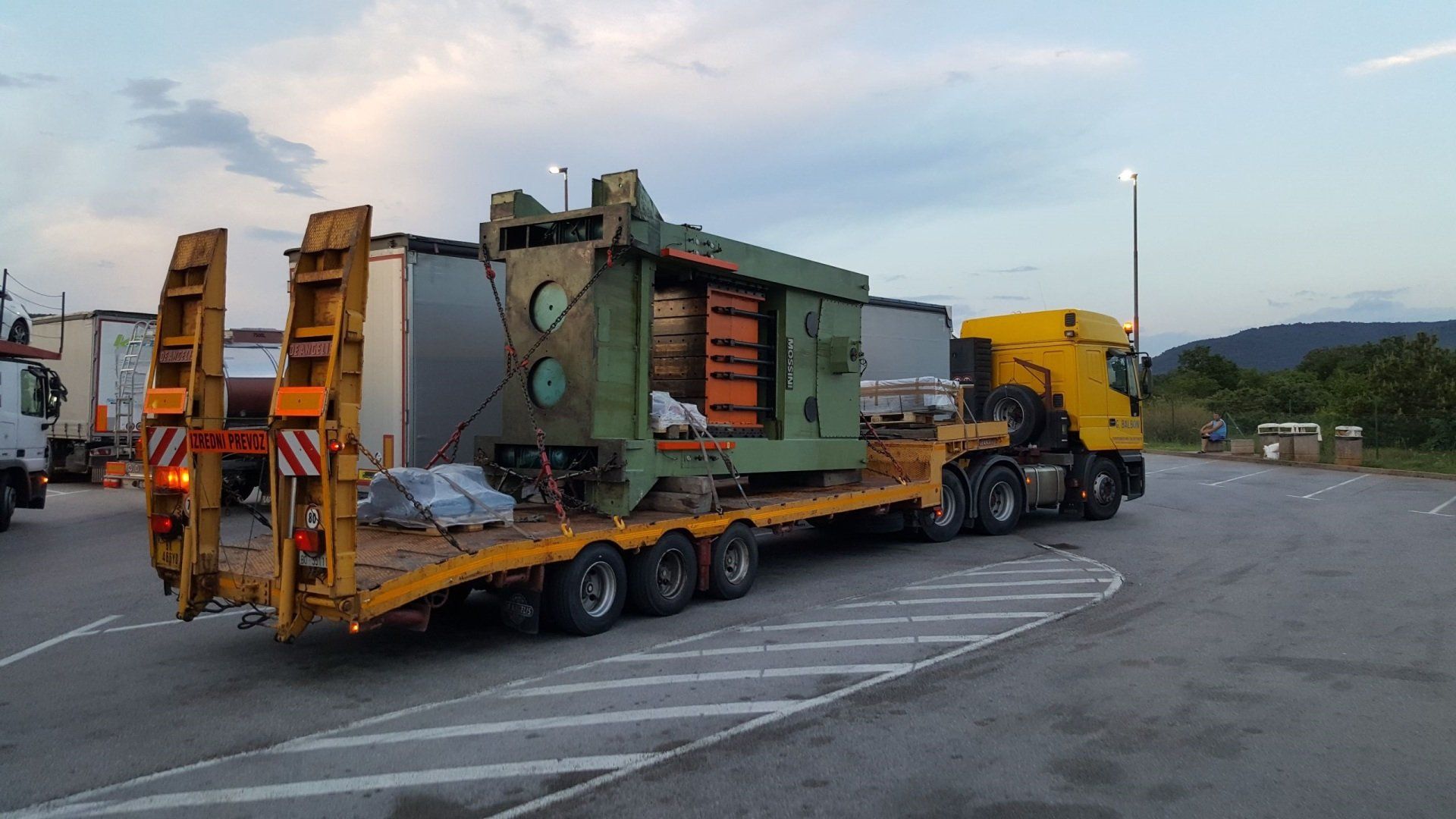 Grande macchina industriale su un camion con pianale, taxi giallo, parcheggiato all'aperto.