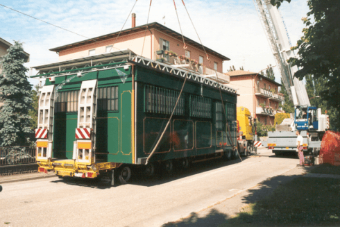 Una grande struttura verde viene sollevata da una gru su un rimorchio a pianale ribassato in una strada con degli edifici.