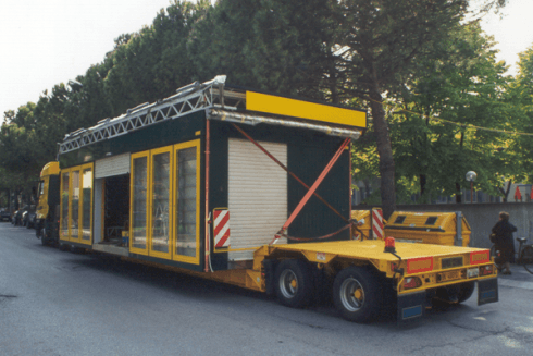 Camion con rimorchio giallo che trasporta un immobile verde scuro e giallo con una persiana bianca.