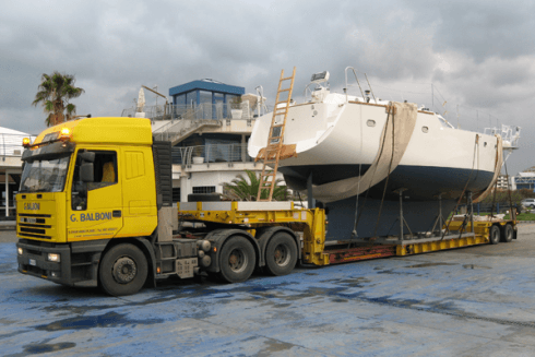 Un camion giallo che trasporta una grande barca a vela bianca e blu su un rimorchio a pianale.