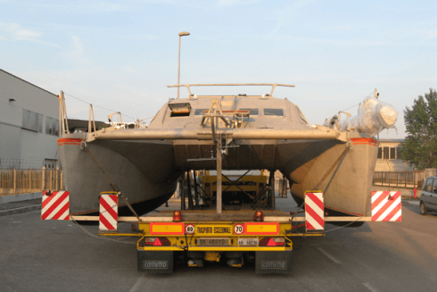 Un catamarano viene trasportato su un rimorchio a pianale, vista posteriore, nei colori grigio e giallo.