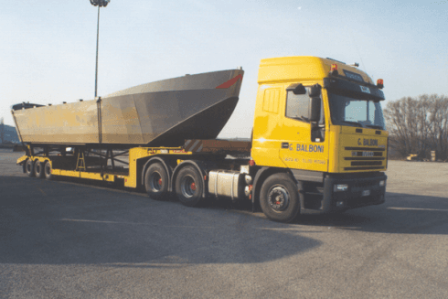 Un semirimorchio giallo con un grande rimorchio grigio per container su un'area asfaltata, sotto un cielo pallido.
