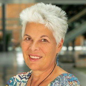 A portrait of a person with short white hair and blue earrings, wearing a patterned top, smiling at the camera.