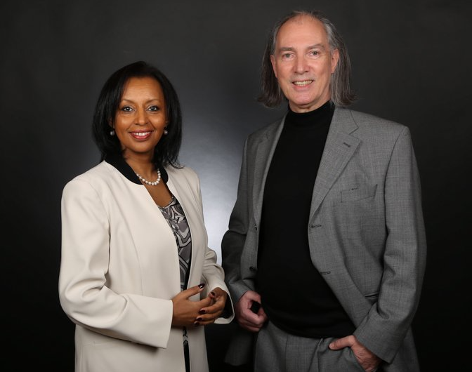 A person in a cream blazer and a person in a grey suit and black turtleneck stand together against a dark background.