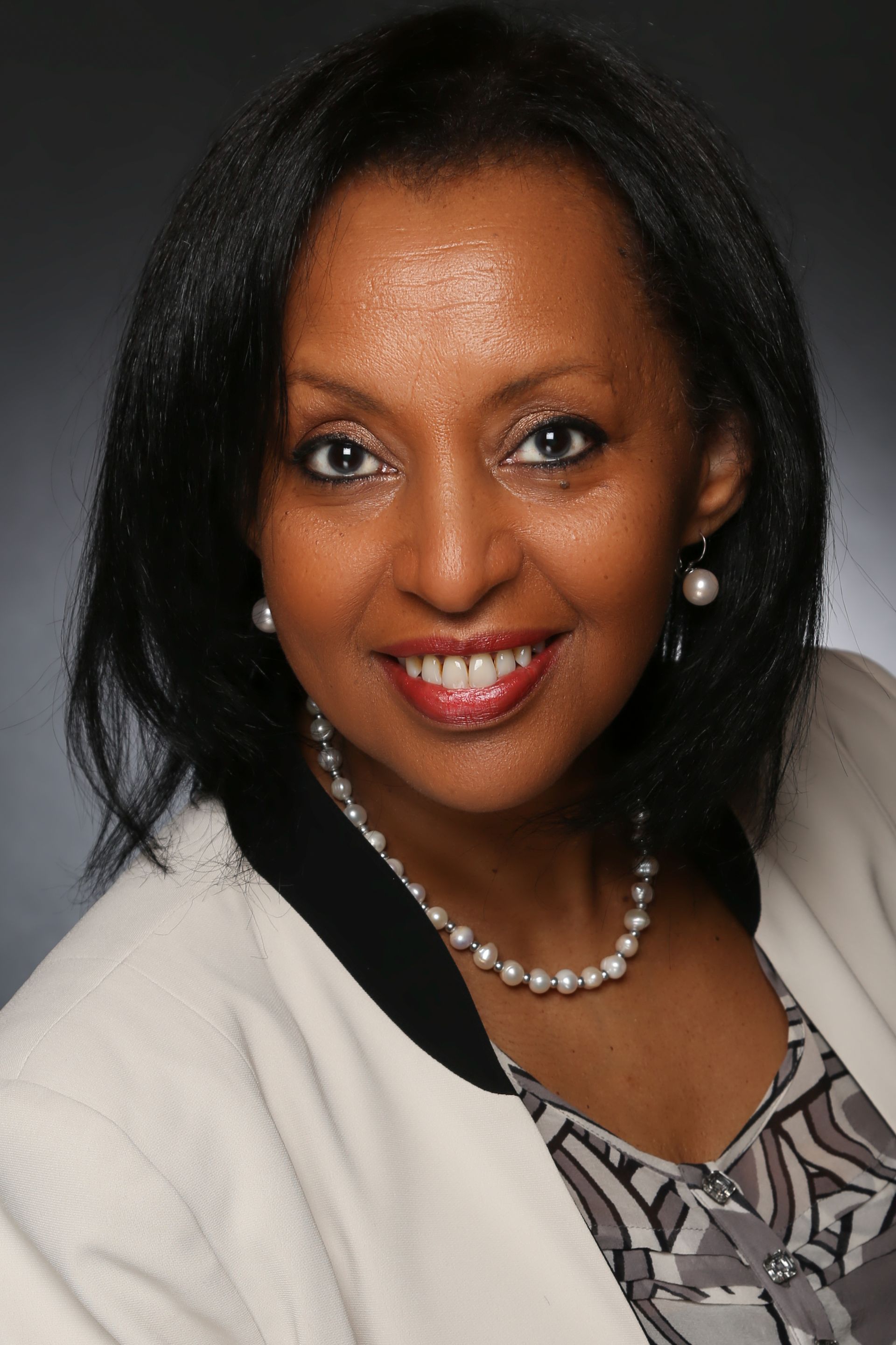 A smiling person in a cream-colored blazer and patterned top, wearing a pearl necklace and drop earrings.
