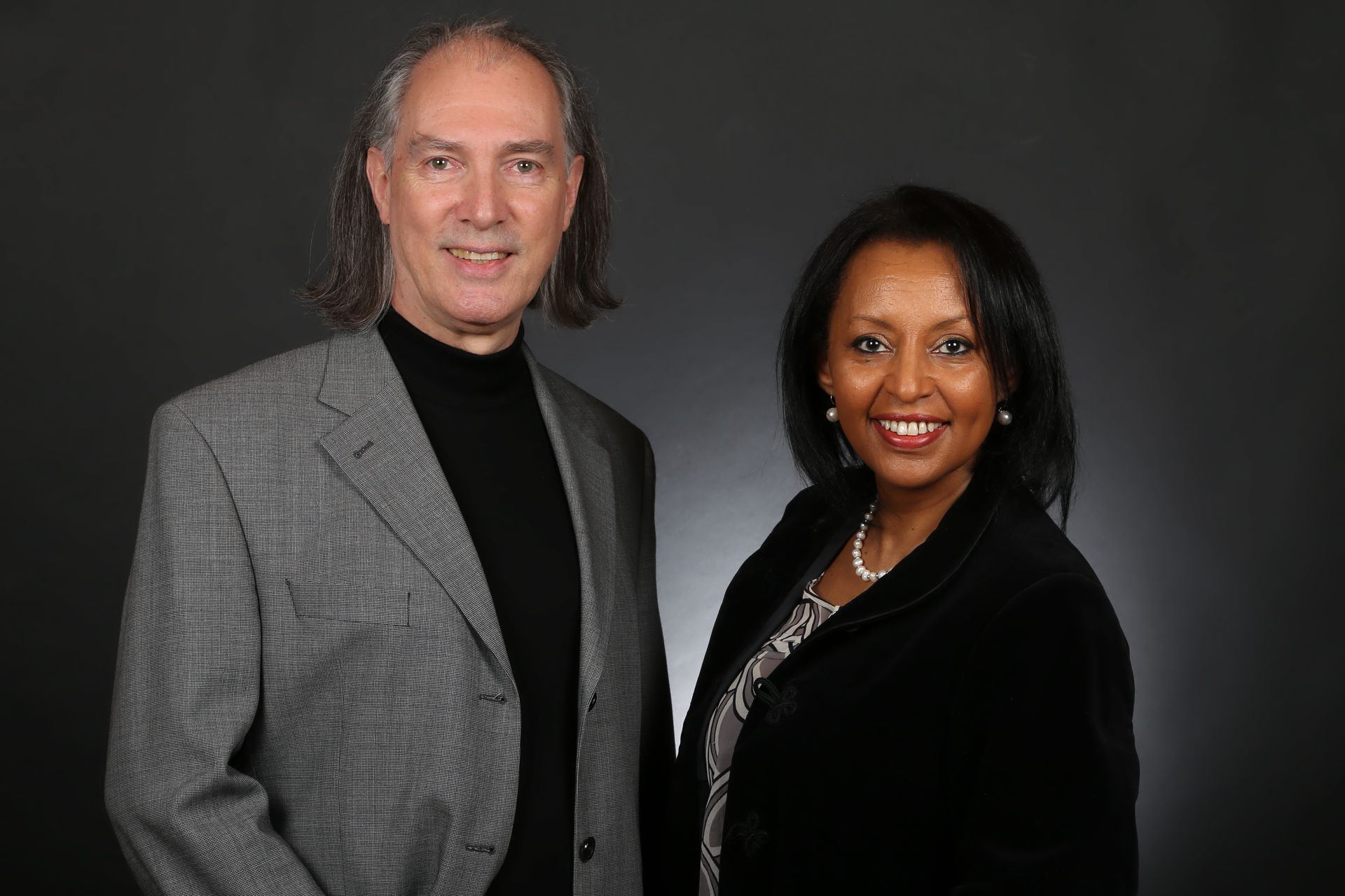 Two professionals standing side-by-side against a dark gray background, smiling at the camera.