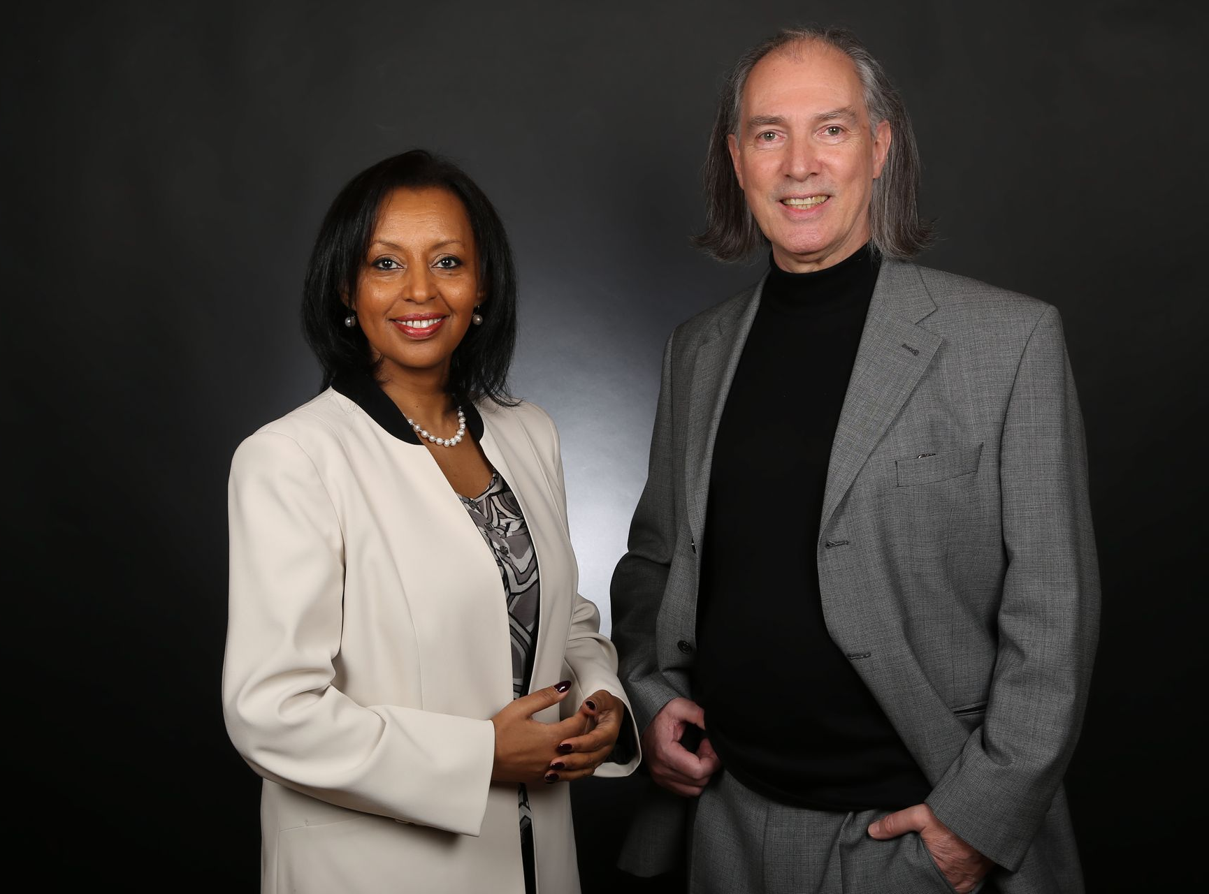 A smiling woman in a white blazer and a man in a grey suit and black turtleneck stand against a dark, neutral background.